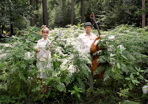 Eine Frau und Mann stehen im Wald umgeben von Farnkraut mit ihren Instrumenten