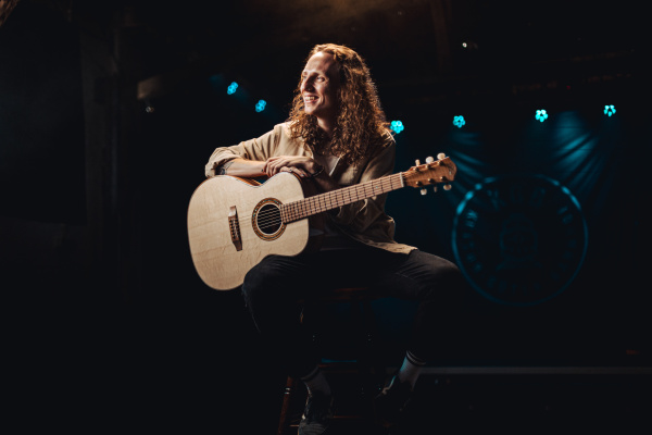Sänger und Songwriter Moe sitzt auf einem Hocker und hält eine helle Akustikgitarre, umgeben von Büh