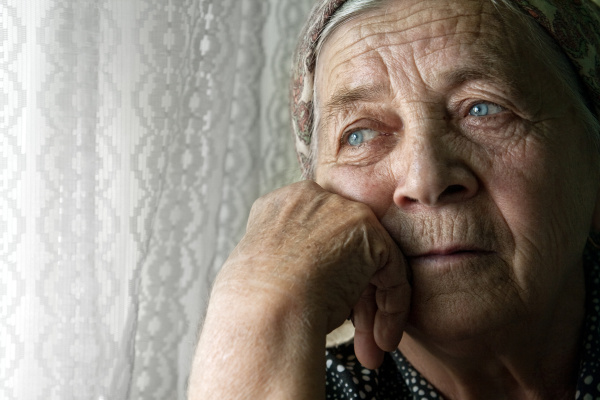 Eine alte Frau mit Kopftuch, die ihren Kopf auf die Hand stützt und traurig aus dem Fenster schaut.