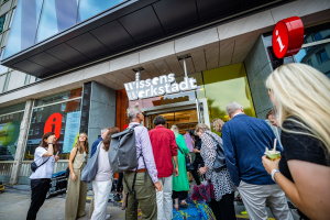 Eine Menschengruppe steht vor dem Eingang der „Wissenswerkstadt Bielefeld“ und betritt das Gebäude. Über dem Eingang hängt das leuchtende Logo, daneben ist die Tourist-Information erkennbar. Auf dem Boden liegen bunte Papierschlangen, einige Personen halten Eisbecher oder Regenschirme.
